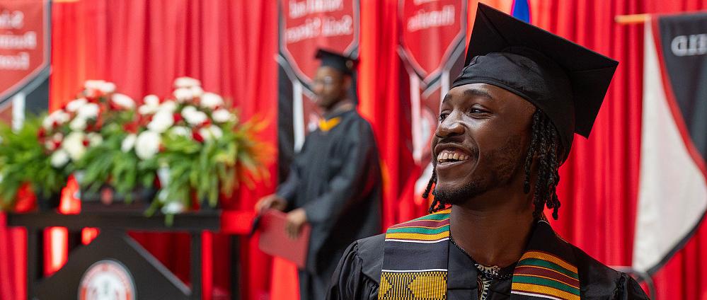 A student in their graduation robes smiling in front a the stage.